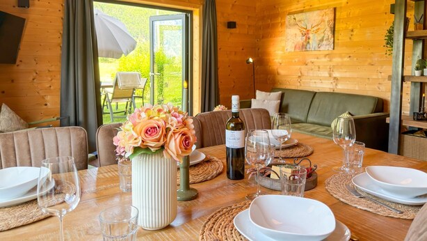 Interior de uma cabana de madeira com uma mesa de jantar posta para dois, um sofá e uma janela grande.