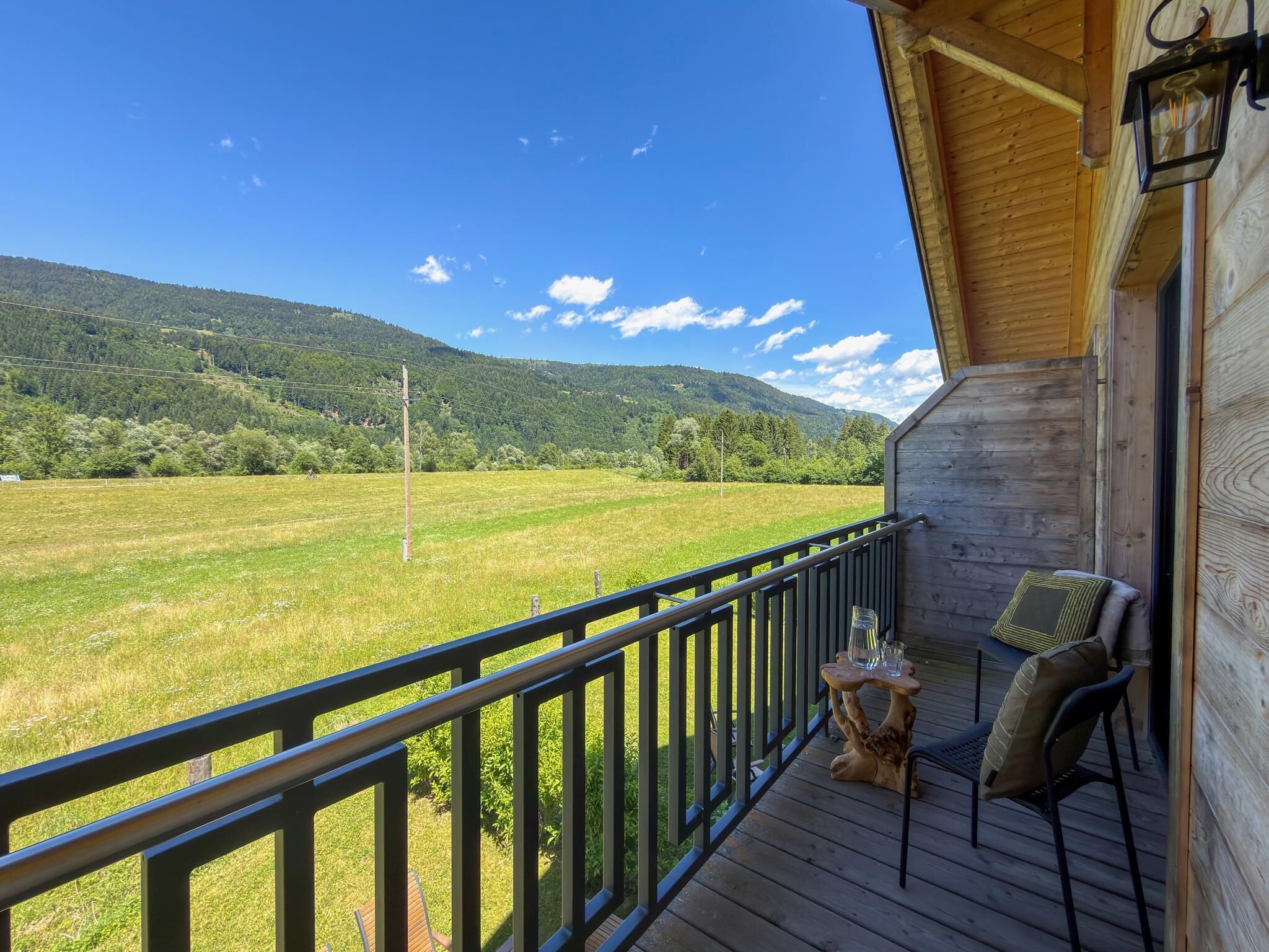 Eine Holzterrasse mit Geländer, einem Tisch mit Stühlen und Blick auf ein grünes Feld und Berge.