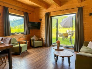 Ein Interieur einer Holzhütte mit Essbereich, einer Couch, einem Fernseher und Blick auf eine grüne Landschaft.