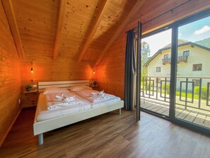 A wooden cabin bedroom with a bed, bedside tables, and a view of a balcony and building outside. - Lovely holiday home on park near ski resort (Rattendorf)