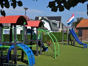 A playground with various play structures, including slides and climbing areas, set against a backdrop of residential buildings and a flag.