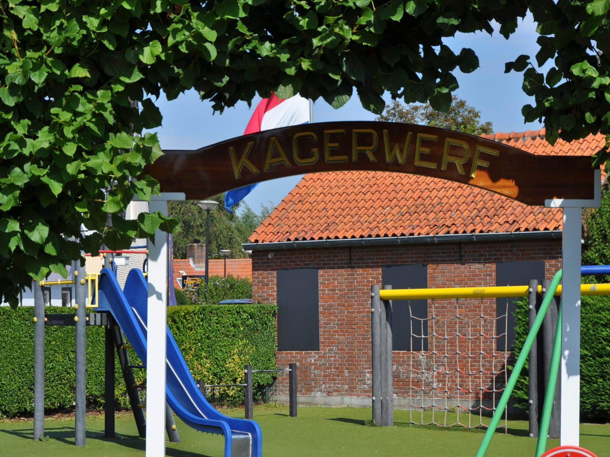 Un parque infantil con un tobogán y estructuras para escalar, un edificio de ladrillo con techo de tejas, y un letrero que dice 'KAGERWERF'.