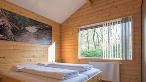 A wooden cabin bedroom with two beds, a large window with a view of trees, and a painting on the wall.