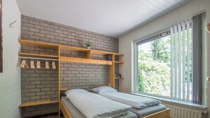 A room with a bed, wooden shelves, a brick wall, and a window with blinds. - Charming single-storey bungalow for 3. (Heesch)