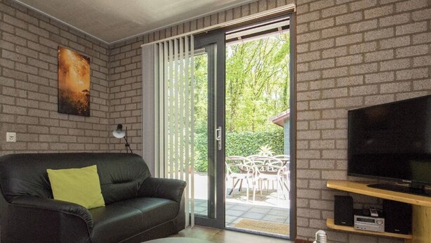 A living room with a black leather sofa, a glass coffee table, a television on a wooden stand, and a sliding glass door leading to an outdoor area with a table and chairs. - Charming single-storey bungalow for 3. (Heesch)
