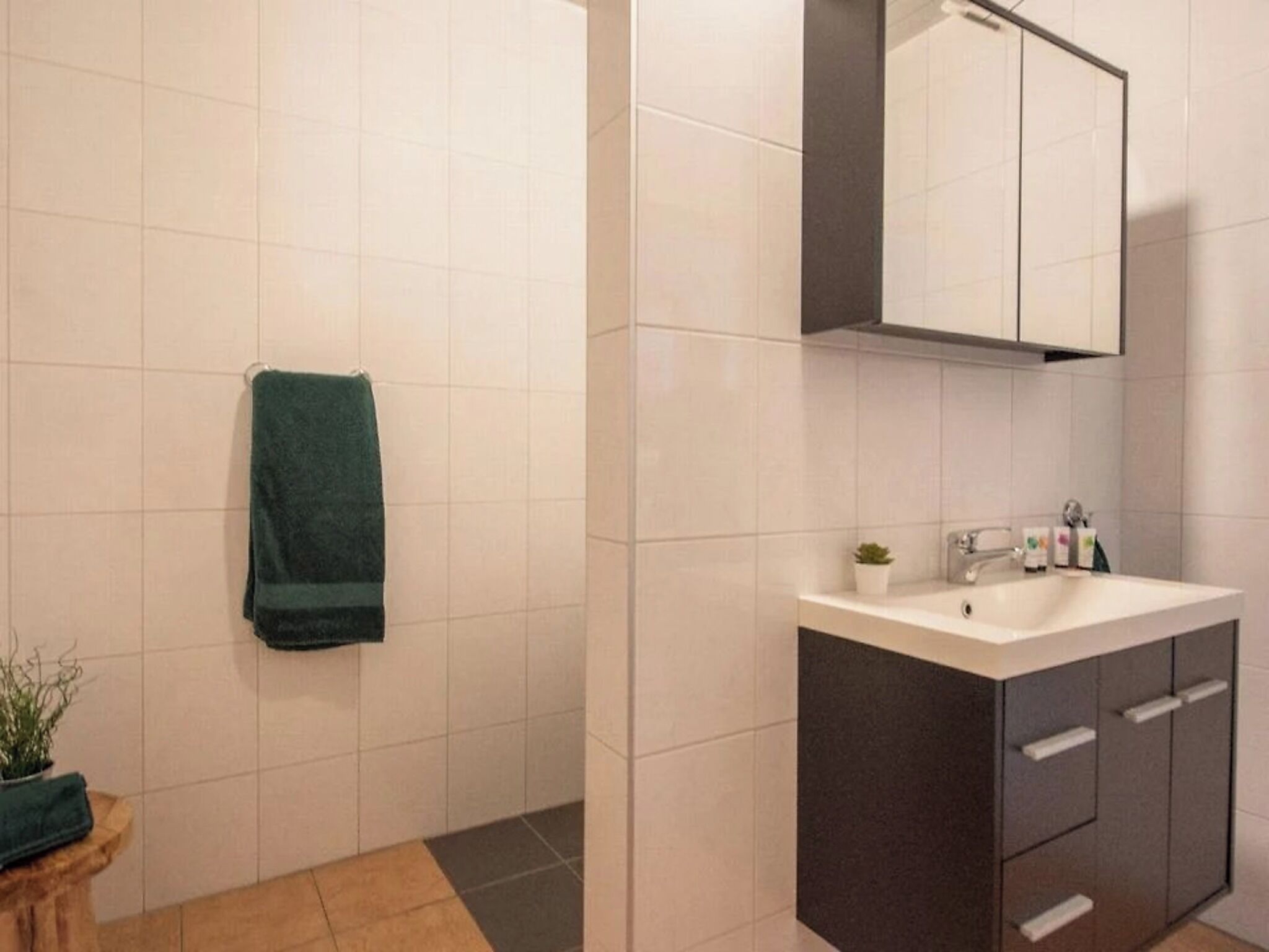 Un baño con azulejos blancos, un mueble oscuro, un lavamanos y una toalla verde colgada en la pared.
