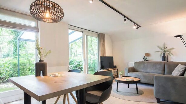 A modern living room with a dining area, a sofa, a TV, and a large window. - Detached lodge with pond view for 4 (Heesch)