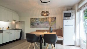 A modern kitchen with white cabinets, a dining table with two chairs, a hanging light fixture, and a large window. - Detached lodge with pond view for 4 (Heesch)