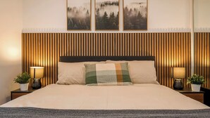 Una cama con cabecera de madera, dos mesitas de noche con lámparas, y tres cuadros enmarcados en la pared.