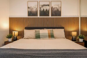 Ein Bett mit Holzkopfteil, zwei Nachttische mit Lampen und drei gerahmte Bilder an der Wand.