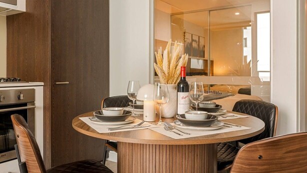Un área de comedor con una mesa redonda, sillas, una vela, copas de vino y una botella de vino.