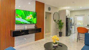 Una sala moderna con un televisor de pantalla plana, un mueble de entretenimiento negro, una mesa redonda con un jarrón de flores y una zona de cocina con un refrigerador y una silla de comedor.