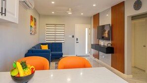 Una sala moderna con un sofá azul, sillones naranja, una mesa de comedor con frutero, un ventilador de techo, iluminación empotrada, un televisor montado en un panel de madera y una puerta con ventanal a rayas.