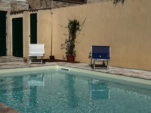 En swimmingpool med två solstolar, en krukväxt och en vägg med en dörr.