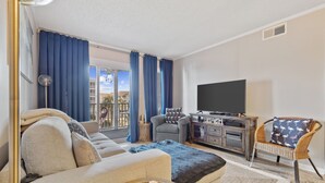 A living room with a sofa, a TV, a wicker chair, and a balcony with blue curtains. - Shore Drive Gem: Walk to Beach, Pool & Apache Pier (Myrtle Beach)