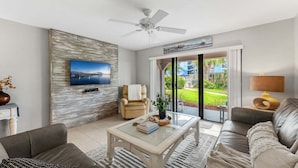 Ein modernes Wohnzimmer mit einem Flachbildfernseher, einem Deckenventilator, einer Couch, einem Couchtisch und Blick auf einen Garten durch Glastüren.