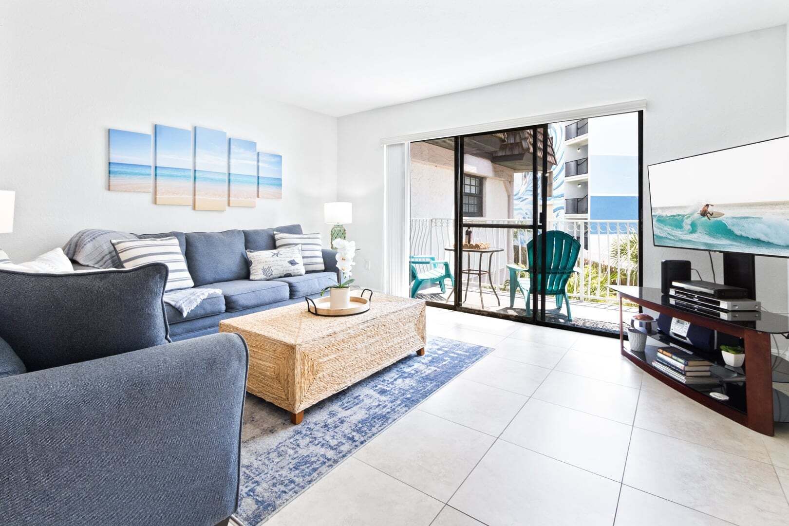 A modern living room with a sofa, coffee table, and a TV. There is a balcony with chairs and a view of the ocean.