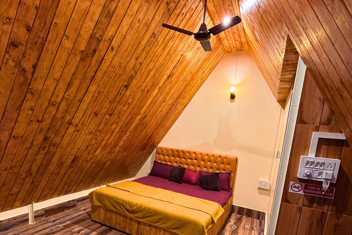 Una habitación con techo inclinado, una cama con colcha amarilla, un ventilador de techo y un cartel de 'No Fumar'.