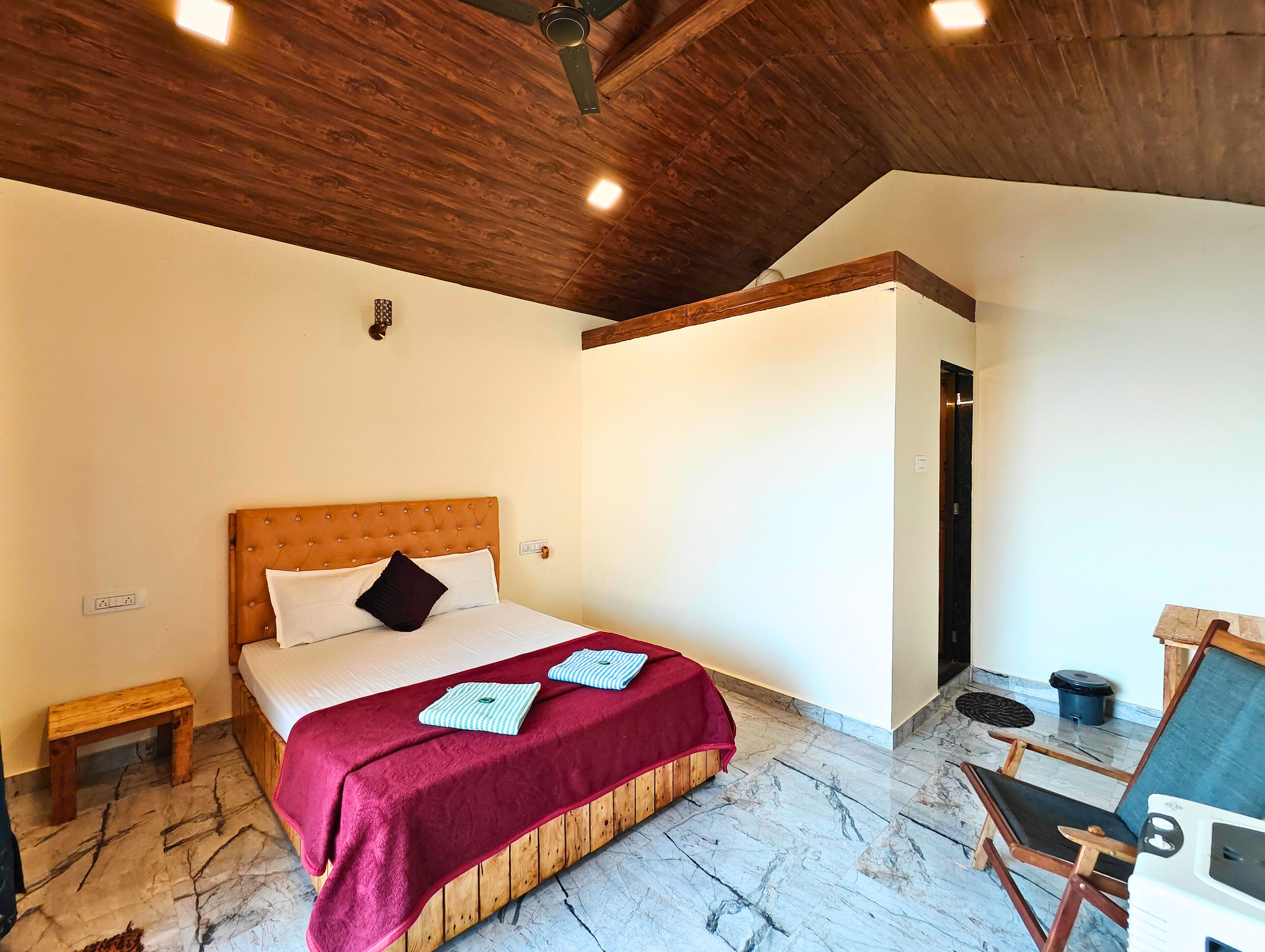 A bedroom with a wooden headboard, a bed with a red blanket, a small wooden stool, and a wooden chair.