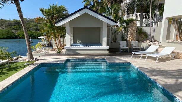 A swimming pool with clear blue water, surrounded by a patio area with lounge chairs and a small building structure. - Romantic Waterfront Villa for 6 (Willemstad)