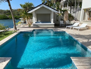 A swimming pool with clear blue water, surrounded by a patio area with lounge chairs and a small building structure. - Romantic Waterfront Villa for 6 (Willemstad)