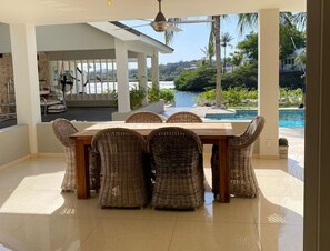 A dining area with a table and wicker chairs, a ceiling fan, and a view of a swimming pool and greenery. - Romantic Waterfront Villa for 6 (Willemstad)