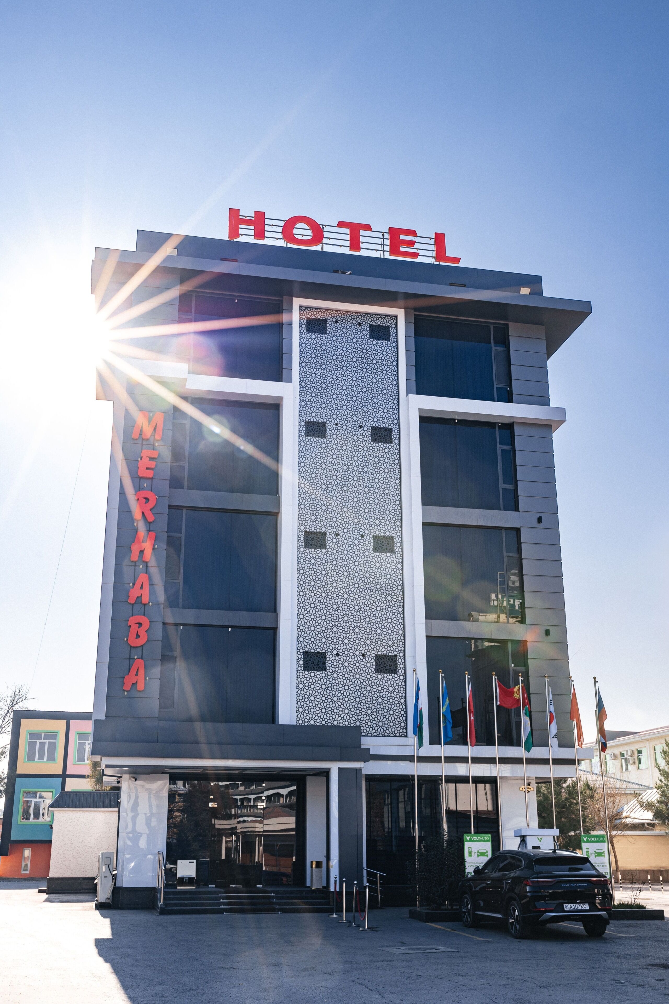 A hotel with a modern design, featuring a prominent 'HOTEL' sign at the top, a vertical sign with 'MERHABA' on the side, and multiple flags displayed outside.