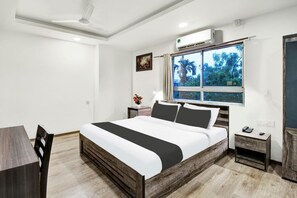 A bedroom with a bed, a desk, a chair, a nightstand, a window with a view, and a ceiling fan. - Super Townhouse Hatigaon (Guwahati)