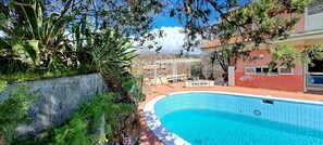 A swimming pool surrounded by greenery, a white fence, and a building with a red roof.