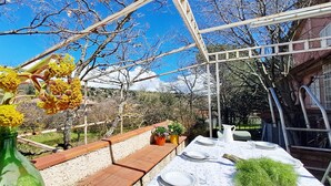 En udendørs spiseplads med et bord dækket til to personer, omgivet af en pergola, en træbænk og plantekasser.