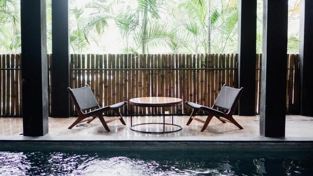 A poolside area with two chairs and a table, bamboo fencing, and a view of palm trees. - Escotel Krisnantara Canggu (Tibubeneng)