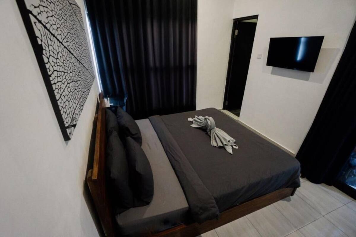 Un lit simple avec un matelas foncé, une tête de lit en bois, un téléviseur à écran plat fixé au mur et une œuvre d’art décorative accrochée au mur.