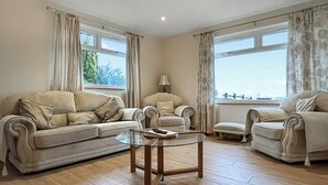 Ein Wohnzimmer mit einer Couch, Sesseln, einem Glastisch und großen Fenstern mit Vorhängen.