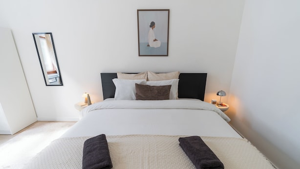 A neatly made bed with pillows, a bedside table with a lamp, a framed picture on the wall, and a mirror on the wall. - Central Ghent Stylish 1BR&1LR Apt WIFI (Ghent)