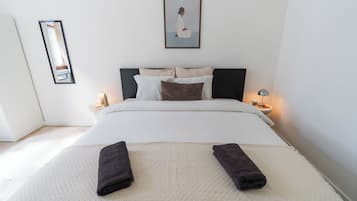Una cama bien hecha con almohadas, una mesita de noche con una lámpara, un cuadro en la pared y un espejo.
