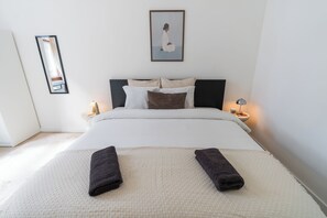 Ein ordentlich bezogenes Bett mit Kissen, ein Nachttisch mit Lampe, ein gerahmtes Bild an der Wand und ein Spiegel.