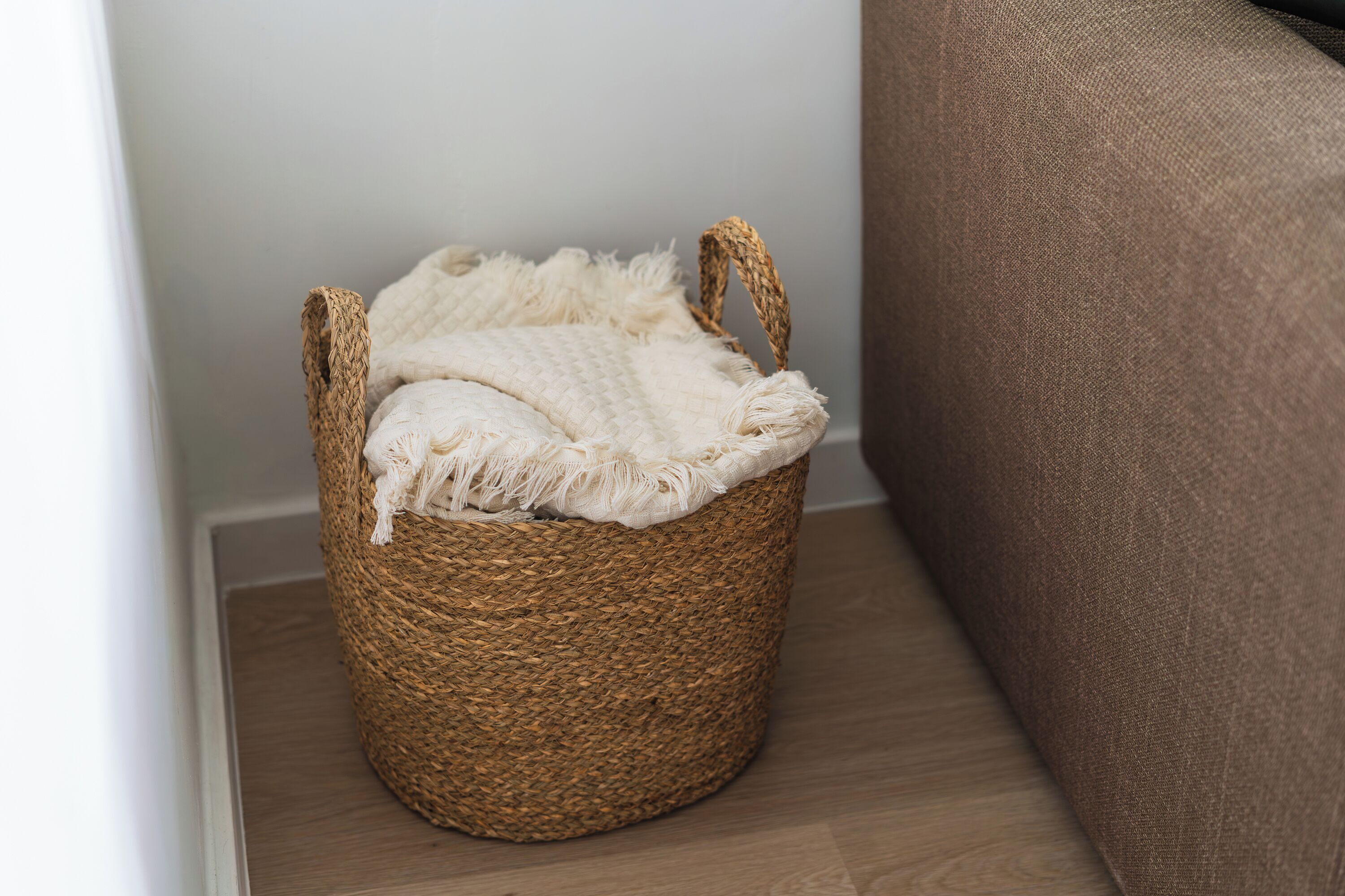 Una cesta tejida con asas, colocada sobre un suelo de madera junto a un mueble tapizado marrón.