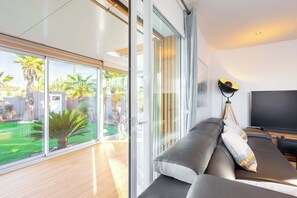 A modern living room with a sofa, a TV, and a large sliding glass door leading to a garden. - Villa Anja Agde Heated Pool & Jacuzzi  Family Villa (Agde)