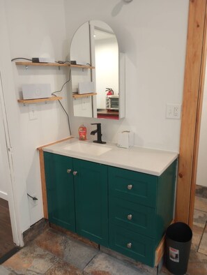 A bathroom with a white countertop, teal cabinets, a black faucet, a mirror, and a trash can.
