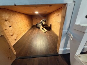 A wooden doghouse with a metal door, a light fixture on the ceiling, and a wooden floor.