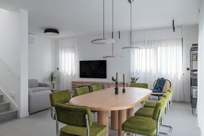 Un comedor moderno con una mesa redonda, sillas tapizadas de verde, luces colgantes, un sofá y un televisor de pantalla plana.