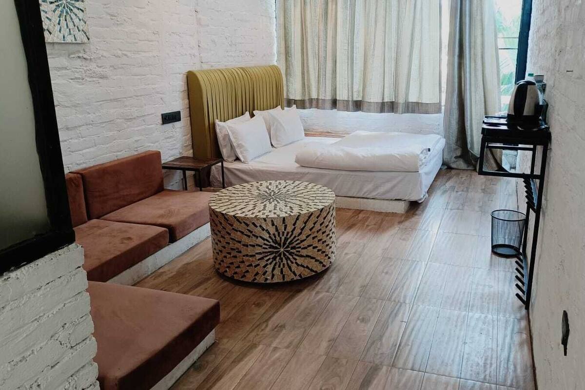 Een kamer met een bed, een slaapbank, een rond poef en een tafeltje met een rode beker.