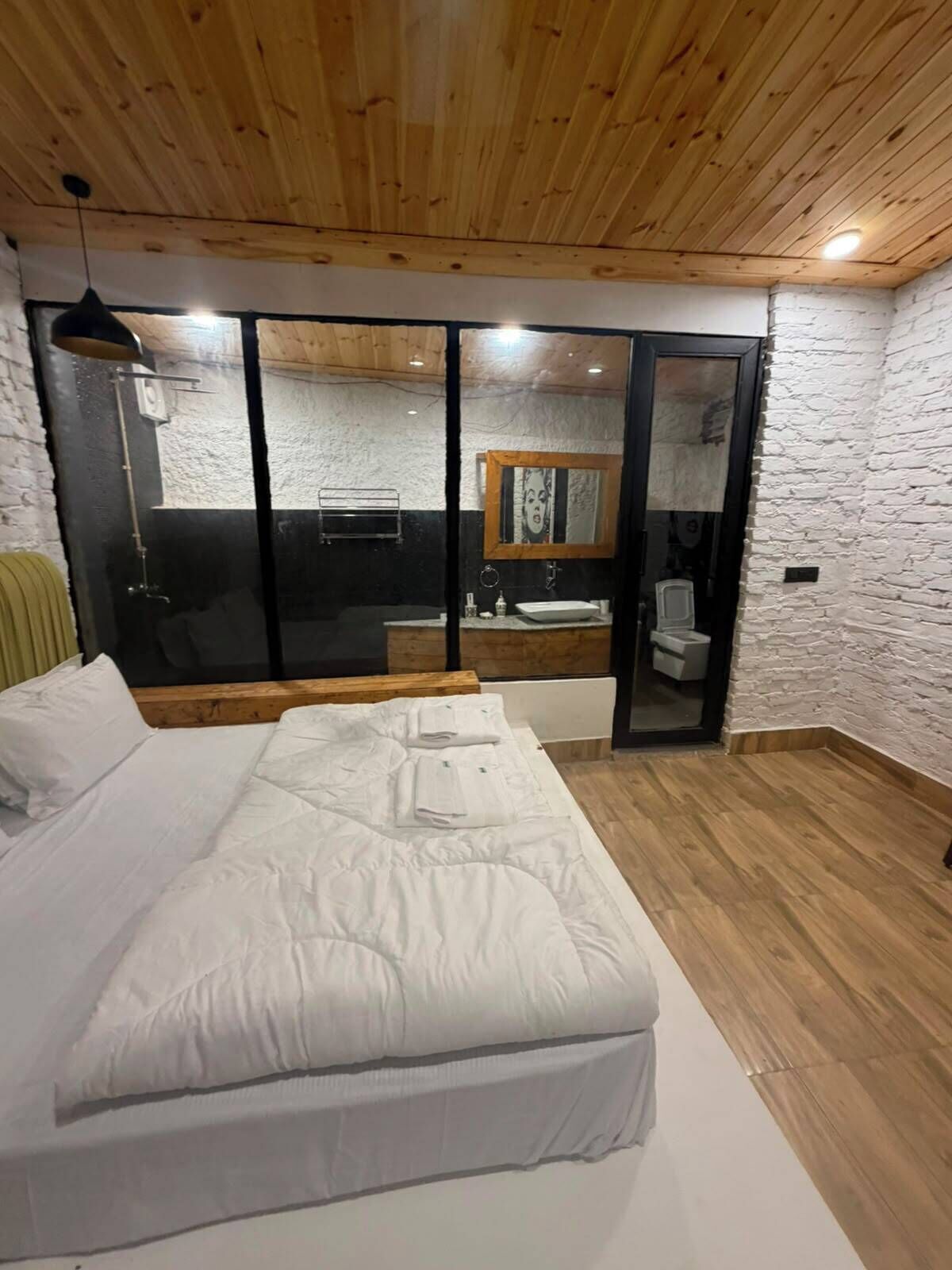 Un dormitorio con una cama, techo de madera, pared de piedra y un baño visible a través de una puerta de cristal.