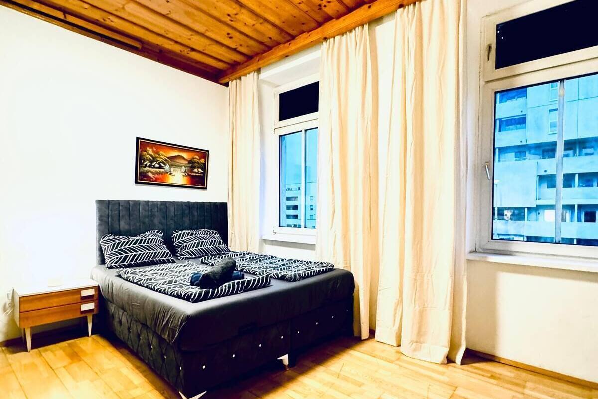 A bedroom with a bed, wooden flooring, a ceiling light, a window with curtains, and a painting on the wall.