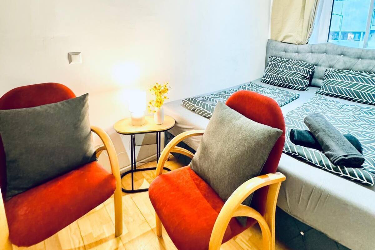 A cozy room with a bed, two chairs, a small table with a lamp, and a window with curtains.
