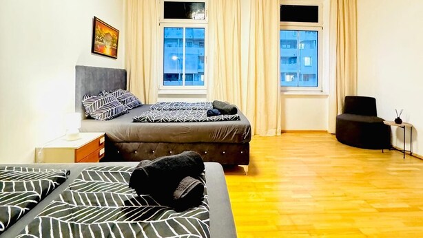 A room with a wooden ceiling, two windows with curtains, a bed with patterned bedding, a chair, and a small table. - Spacious Private Room Near FAC Platz (Wien)