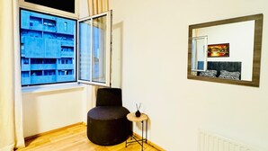 A room with wooden flooring, a black ottoman, a small table with a candle, a window with a view of a building, and a radiator. - Spacious Private Room Near FAC Platz (Wien)