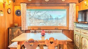 Ein Esszimmer aus Holz mit einem großen Fenster, einem Tisch, Stühlen, einem Heizkörper und einem Fernseher.
