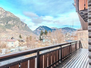 Ein Balkon mit Blick auf eine gebirgige Landschaft, ein Dorf weiter unten und ein Holzhaus zur Rechten.