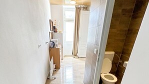 Un couloir étroit, dont le sol est recouvert de carreaux, mène à une salle de bain équipée de toilettes et d’un espace douche.